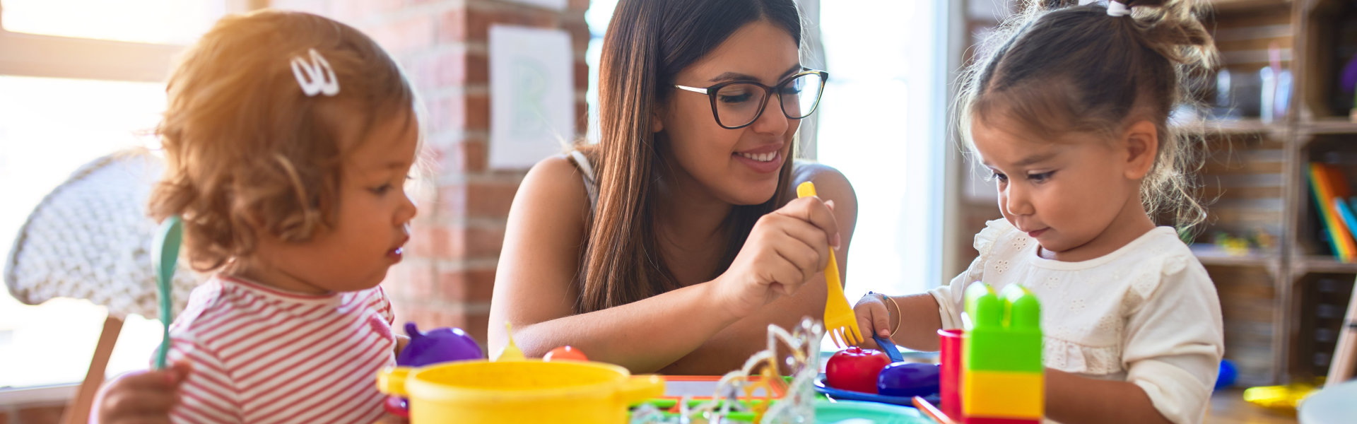 Young beautiful teacher and toddlers playing meals using plastic food and cutlery toy at kindergarten