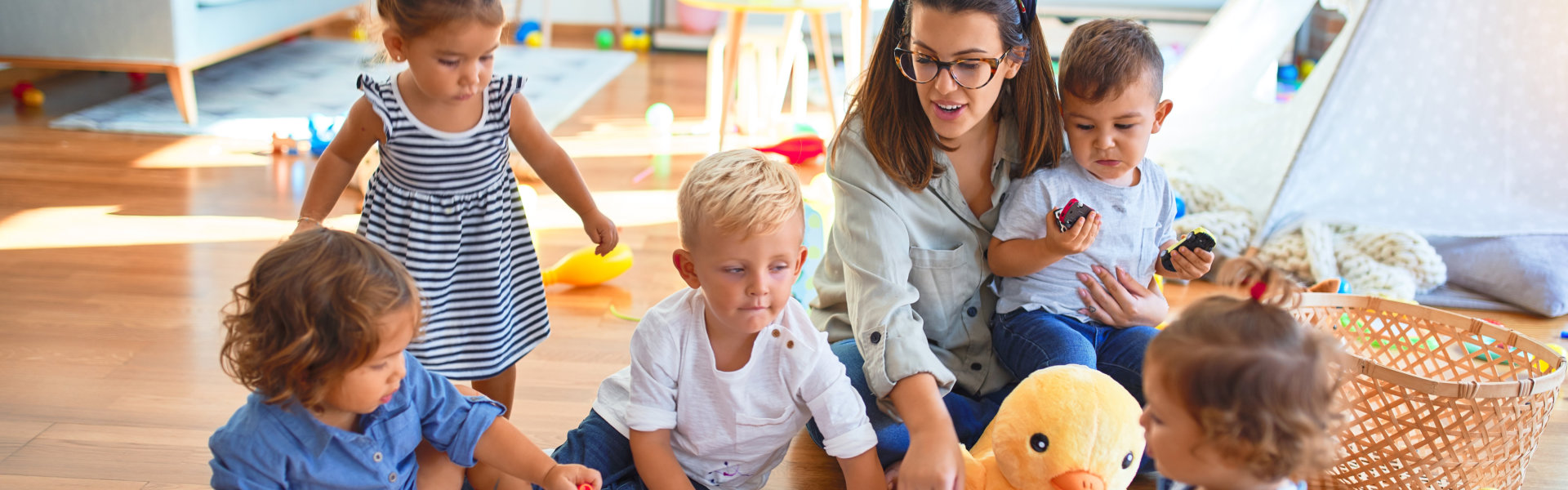 Beautiful teacher and group of toddlers sitting on the floor drawing using paper and pencil around lots of toys at kindergarten